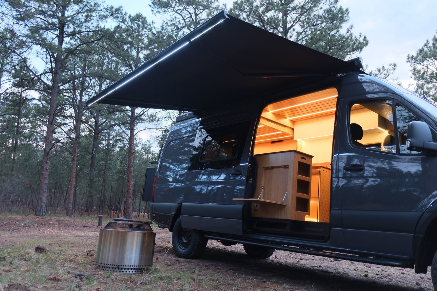 The Den parked in a pine forest with the awning out