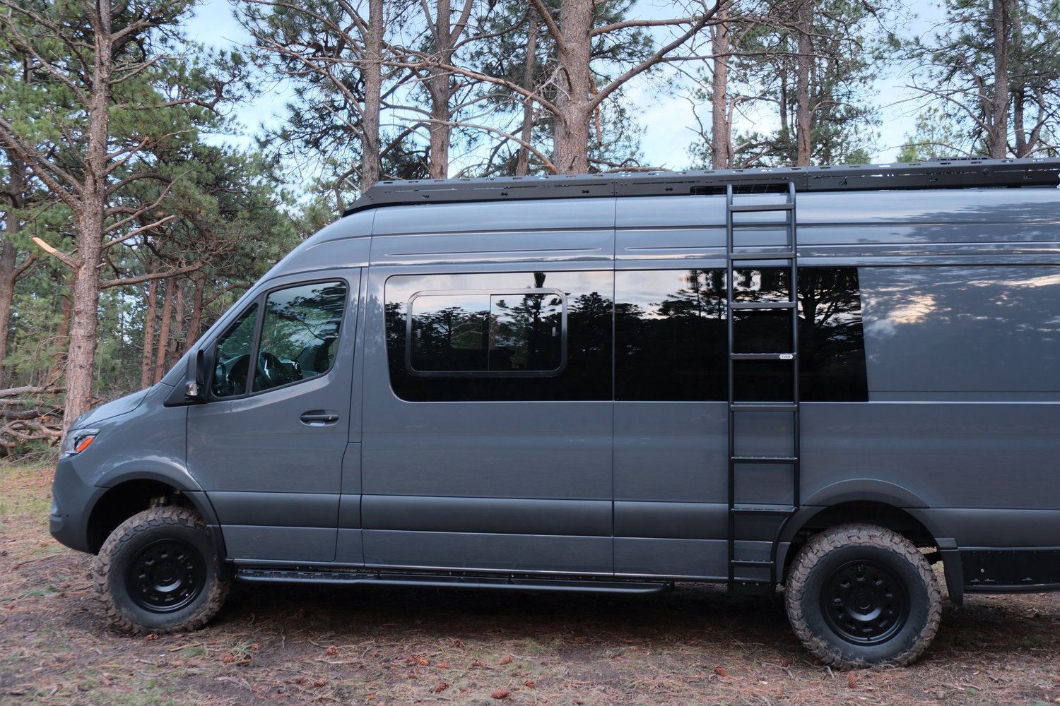 Side view of the Den gray Sprinter camper parked among pine trees.