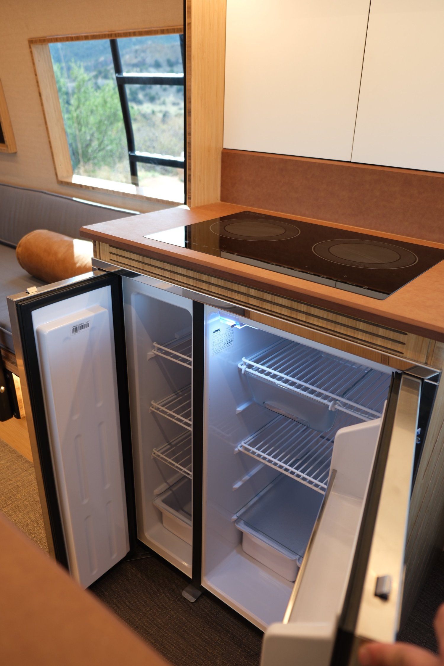 The Den refrigerator with both doors open beneath a wood-toned countertop and induction cooktop.