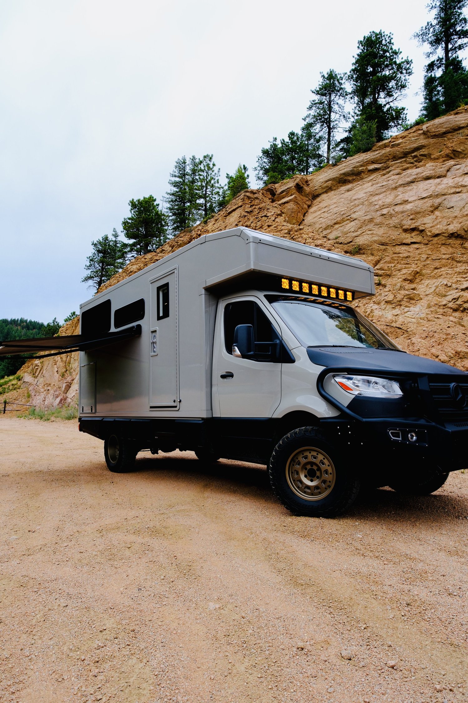 The Nest composite-body adventure vehicle beside a rocky, pine-covered hillside.